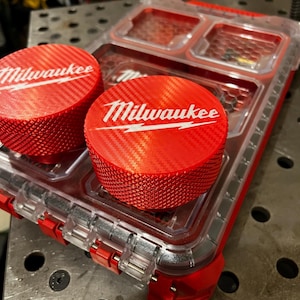 May include: A red and clear plastic storage container with two red, textured, circular lids. The lids have the white "Milwaukee" logo. The container has a clear top with two compartments and red accents.