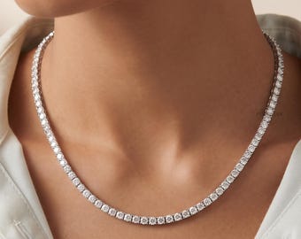 May include: A silver-toned tennis necklace featuring a continuous row of round, clear, faceted stones set in a metallic chain. The necklace rests around the neck, worn over a white collared shirt with visible buttons and a textured fabric finish.