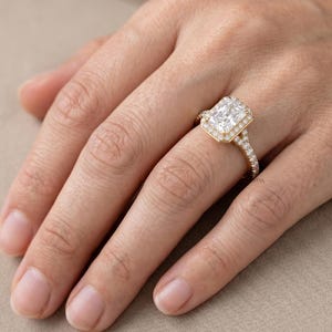 Può includere: Un anello d'oro con una grande pietra preziosa trasparente rettangolare al centro, circondata da pietre preziose trasparenti più piccole. Anche la fascia dell'anello è ornata da piccole pietre preziose trasparenti. L'anello è su un dito.