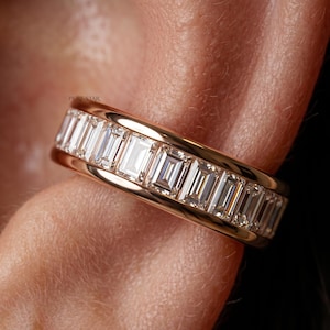 Può includere: Un anello a fascia in oro con una fila continua di pietre preziose rettangolari e trasparenti. L'anello è incastonato con più pietre taglio baguette, creando un effetto scintillante. La foto ravvicinata mette in risalto il design dell'anello e la brillantezza delle pietre.