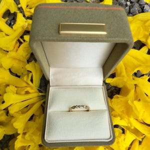 Könnte beinhalten: Ein goldener Ring mit mehreren kleinen Steinen wird in einer offenen, olivgrünen Schmuckschatulle mit einem goldenen Namensschild präsentiert. Die Schatulle ist mit cremefarbenem Stoff ausgekleidet. Der Hintergrund ist mit gelben Blütenblättern bedeckt.