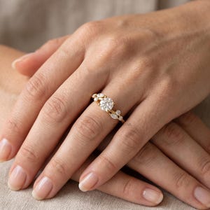 May include: A gold ring featuring a large round brilliant-cut centre stone flanked by smaller marquise and round-cut stones arranged in a cluster along the band. The metal band is polished gold, and the stones are clear and faceted.