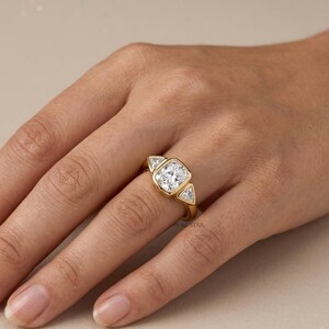 Puede incluir: Un anillo de oro con una piedra preciosa central transparente de corte coj&iacute;n en un engaste de bisel, flanqueada por dos piedras transparentes m&aacute;s peque&ntilde;as de corte triangular a cada lado. La banda es de oro amarillo pulido con tres piedras.