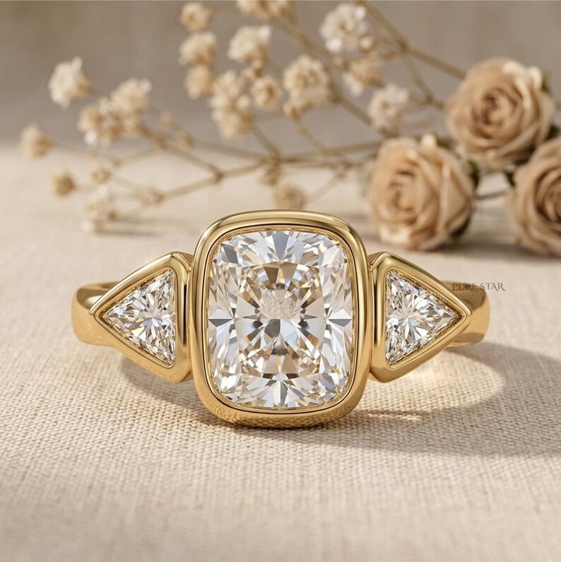 Puede incluir: Un anillo de oro con una gran piedra central de corte coj&iacute;n en un engaste de bisel, flanqueada por dos piedras m&aacute;s peque&ntilde;as de corte triangular a cada lado. La banda es de oro pulido y el texto PURE STAR es visible en el lado derecho.