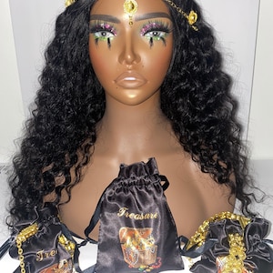 May include: A mannequin head with long, wavy black hair, adorned with a gold headpiece. Three black satin drawstring bags with gold text and a treasure chest graphic are displayed. Gold jewelry is visible.