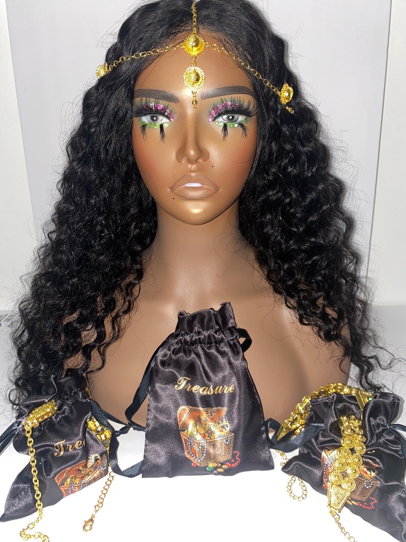May include: A mannequin head with long, wavy black hair, adorned with a gold headpiece. Three black satin drawstring bags with gold text and a treasure chest graphic are displayed. Gold jewelry is visible.