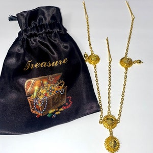 May include: A gold-colored headpiece with a chain design and circular embellishments. A black satin pouch with the word "Treasure" and a treasure chest illustration is next to the headpiece.