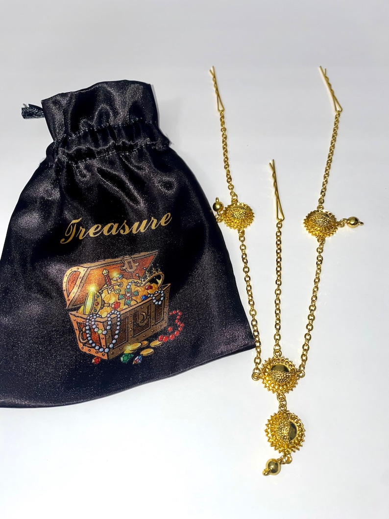 May include: A gold-colored headpiece with a chain design and circular embellishments. A black satin pouch with the word "Treasure" and a treasure chest illustration is next to the headpiece.
