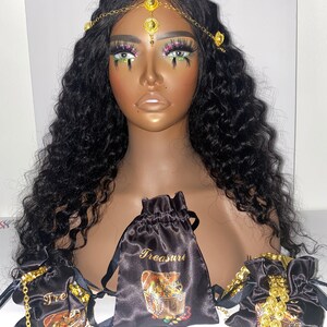 May include: A mannequin head with long, wavy black hair and a gold-toned headpiece. Several black satin drawstring bags with gold text and treasure chest illustrations are displayed. Gold jewelry is visible.