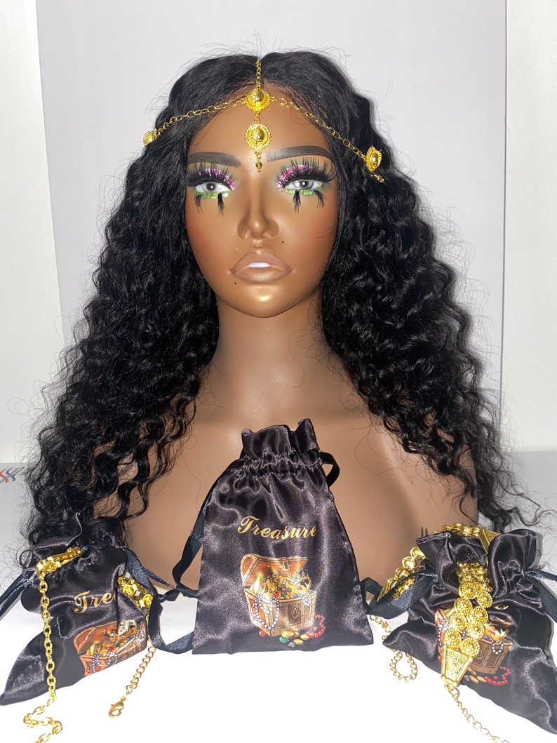 May include: A mannequin head with long, wavy black hair and a gold-toned headpiece. Several black satin drawstring bags with gold text and treasure chest illustrations are displayed. Gold jewelry is visible.