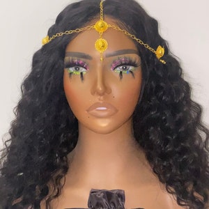 May include: A gold-toned headpiece with ornate circular and teardrop-shaped embellishments is draped across a mannequin's dark hair. A black satin drawstring bag with the word "Treasure" in gold script is visible.