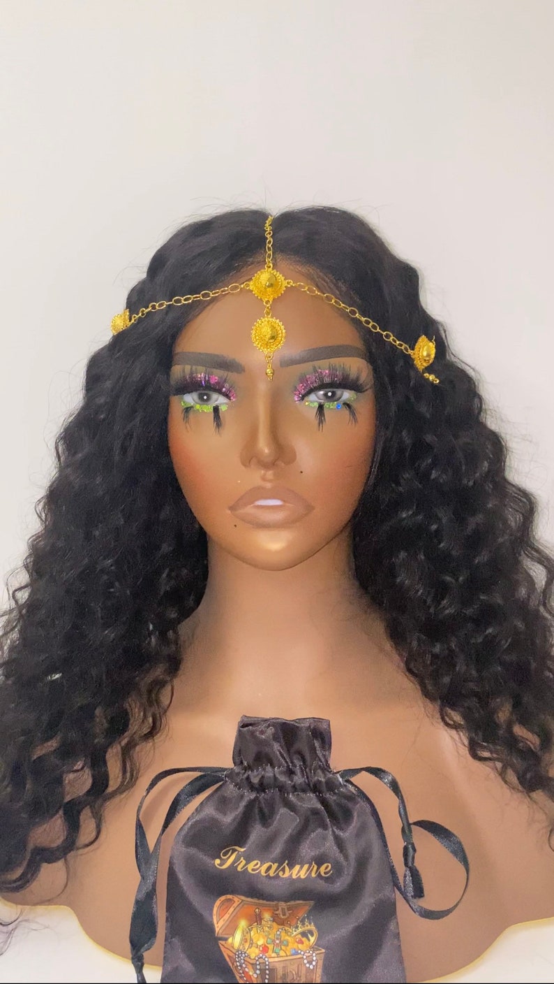 May include: A gold-toned headpiece with ornate circular and teardrop-shaped embellishments is draped across a mannequin's dark hair. A black satin drawstring bag with the word "Treasure" in gold script is visible.