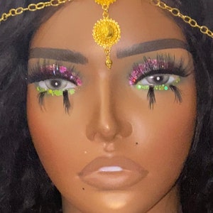 May include: A gold-colored headpiece with a central floral design and chain accents. The model has dramatic eye makeup with pink and green glitter, and long, dark eyelashes. The skin tone is a warm brown.