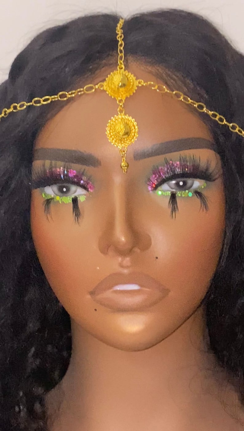 May include: A gold-colored headpiece with a central floral design and chain accents. The model has dramatic eye makeup with pink and green glitter, and long, dark eyelashes. The skin tone is a warm brown.