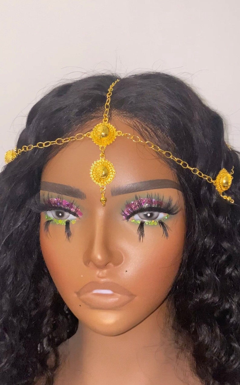 May include: A gold-colored headpiece with a floral design, worn on a person with dark hair. The headpiece is made of linked chains and circular ornaments. The person has dramatic eye makeup with pink and green accents.