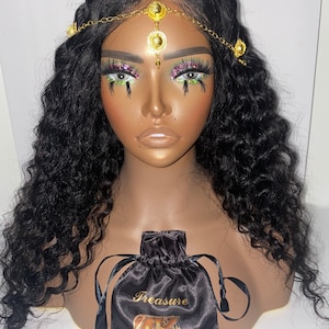 May include: A mannequin head with long, wavy black hair and a gold-colored headpiece. The headpiece has decorative circular accents. A black satin drawstring bag with the word "Treasure" and a treasure chest graphic is in front.