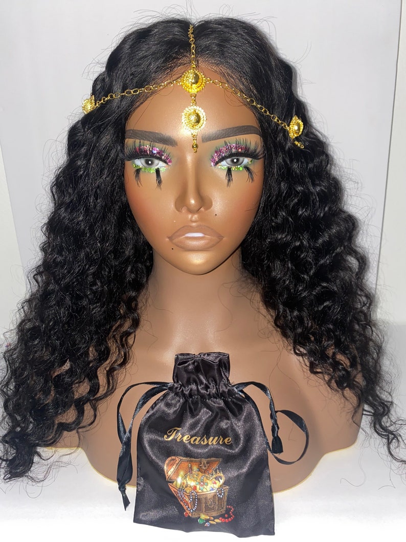 May include: A mannequin head with long, wavy black hair and a gold-colored headpiece. The headpiece has decorative circular accents. A black satin drawstring bag with the word "Treasure" and a treasure chest graphic is in front.