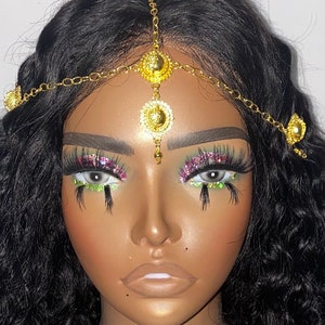 May include: A gold-colored headpiece with a chain and circular pendants. The headpiece is worn on a mannequin head with dark curly hair. The mannequin has dramatic eye makeup with long eyelashes and glitter.