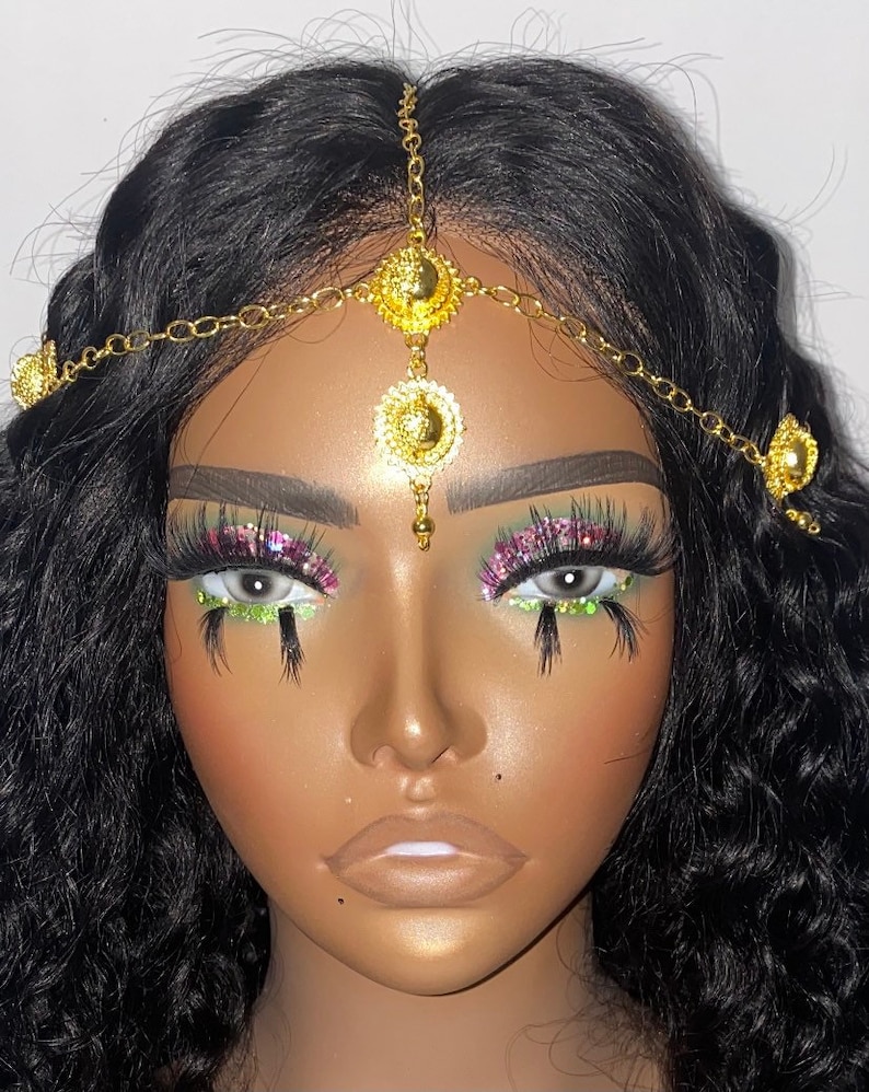 May include: A gold-colored headpiece with a chain and circular pendants. The headpiece is worn on a mannequin head with dark curly hair. The mannequin has dramatic eye makeup with long eyelashes and glitter.
