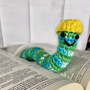 May include: A crocheted green, blue, and white worm with a yellow crocheted hat on top of a book. The book is open and the text "HENRY THE FIFTH" is visible on the page.