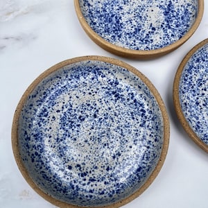 May include: Three ceramic bowls with a speckled blue and white design on the inside. The rims are a natural, unglazed brown. The bowls are round and have a shallow depth, suitable for serving food or as decorative pieces.