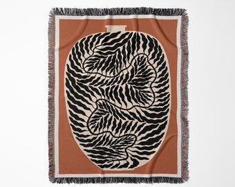 Op de afbeelding: Een geweven sprei met een groot, gestileerd vaasontwerp. De vaas is gevuld met zwart- en crèmekleurige bladvormige patronen. De deken heeft een roestkleurige achtergrond met een crèmekleurige rand en franjes.