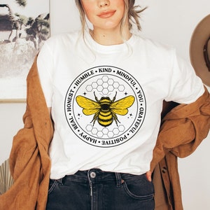 以下が含まれることがあります： 黒と黄色の蜂のグラフィックが描かれた白いTシャツ。蜂は、「Honest、Humble、Kind、Mindful、You、Grateful、Positive、Happy、Real」という文字が書かれた円に囲まれています。