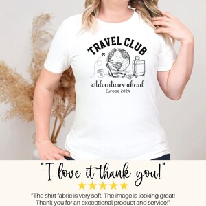 May include: White t-shirt with a black graphic design of a globe, airplane, suitcase, camera, and passport. The text "TRAVEL CLUB" is above the graphic and "Adventures ahead Europe 2024" is below the graphic.