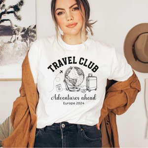 May include: White t-shirt with a black graphic of a globe, suitcase, camera, and passport. The text "Travel Club" is above the graphic and "Adventures ahead Europe 2024" is below.