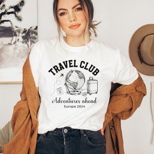 May include: White t-shirt with a black graphic design that says "Travel Club" with a globe, airplane, passport, camera, and suitcase. The text below the graphic says "Adventures ahead Europe 2024".