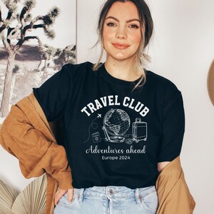 May include: A black t-shirt with a white graphic design that says "Travel Club" with a globe, airplane, suitcase, camera, and passport. The text below the graphic says "Adventures ahead Europe 2024".
