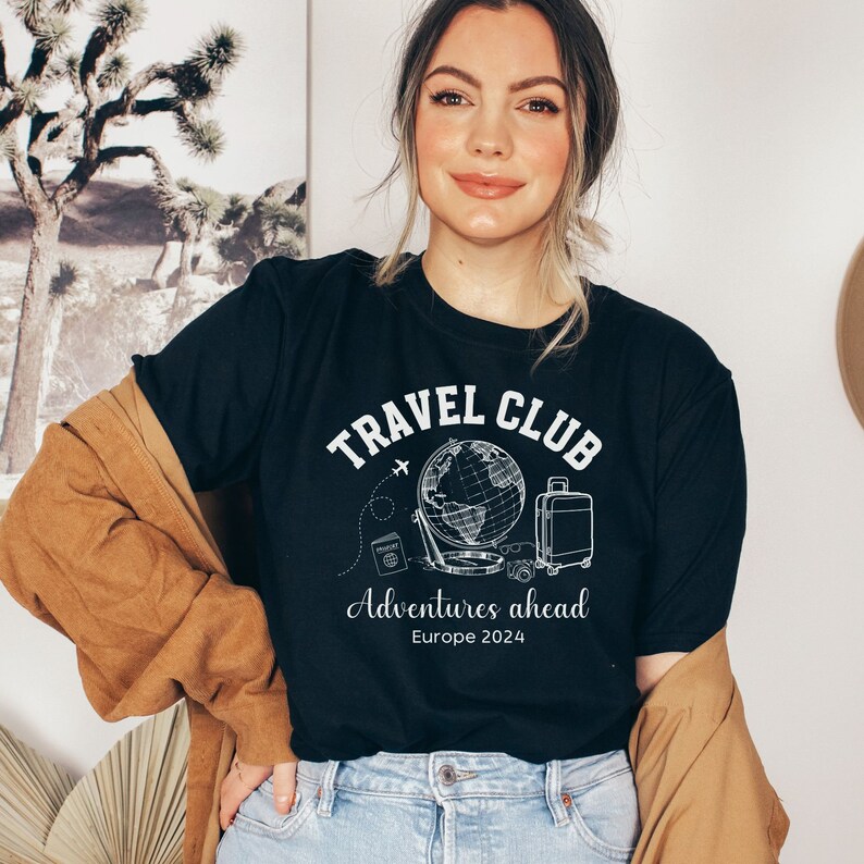 May include: A black t-shirt with a white graphic design that says "Travel Club" with a globe, airplane, suitcase, camera, and passport. The text below the graphic says "Adventures ahead Europe 2024".