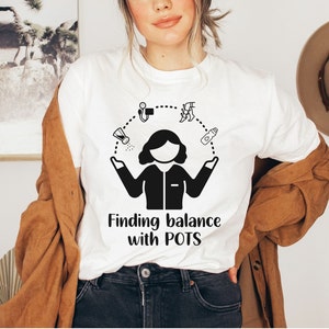 May include: White t-shirt featuring a black graphic of a woman juggling various items, including a water bottle, a salt shaker, a medication bottle, and a heart rate monitor. The text "Finding balance with POTS" is printed below the graphic.