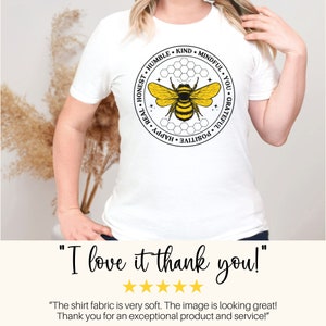 以下が含まれることがあります： 黒と黄色の蜂のグラフィックが描かれた白いTシャツ。蜂は、「正直、謙虚、親切、思いやり、あなた、感謝、前向き、幸せ、本物」という言葉が書かれた円に囲まれています。