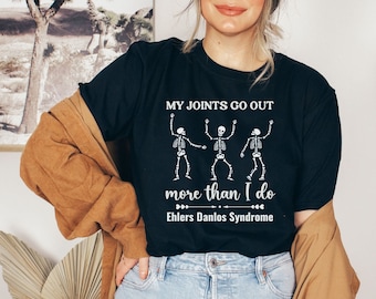 Puede incluir: Camiseta negra con un gráfico blanco de tres esqueletos bailando y el texto "Mis articulaciones se salen más que yo Ehlers Danlos Syndrome".