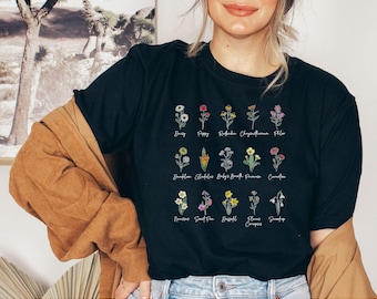 Może przedstawiać: Czarny T-shirt z białym nadrukiem różnych kwiatów i ich nazwami. Kwiaty obejmują stokrotkę, mak, redbud, chryzantemę, bez, paprotkę, mieczyk, gipsówkę, Francję, goździk, narcyza, groszek pachnący, narcyza, dzwonki, śnieżynkę.
