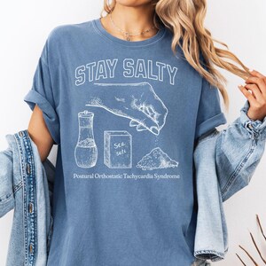 May include: A blue t-shirt featuring the phrase "STAY SALTY" in white. The design includes a hand-drawn illustration of a hand sprinkling salt, a salt shaker, and a box of sea salt. The text "Postural Orthostatic Tachycardia Syndrome" is printed below the illustration.