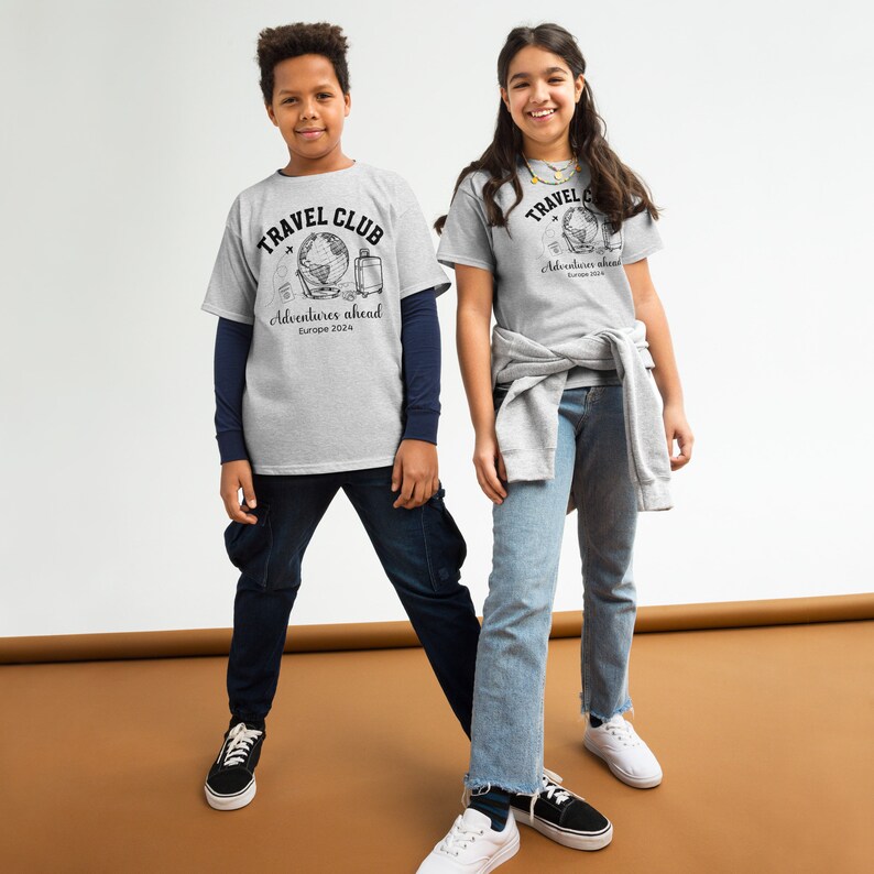 May include: Two kids wearing grey t-shirts with a globe graphic and the text "Travel Club Adventures Ahead Europe 2024". The boy is wearing blue jeans and the girl is wearing light blue jeans.