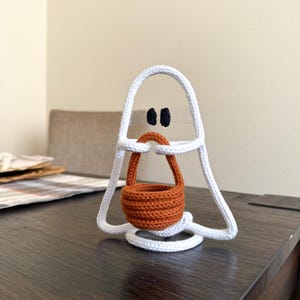 May include: A white crocheted ghost figure with two small black oval eyes, holding a small, round, orange crocheted basket with a handle. The ghost has a triangular shape with a circular base and is constructed from a continuous white cord.