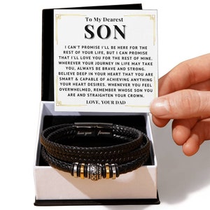 Peut inclure: Un bracelet en cuir tressé noir avec des détails argentés et dorés est présenté dans un coffret cadeau blanc. Une carte avec le texte "To My Dearest SON" et un message est tenue au-dessus du bracelet. Le bracelet porte l'inscription "love you forever".