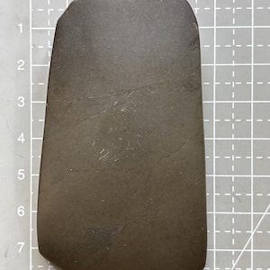 May include: A dark gray, smooth stone axe head with a tapered shape and a flat, slightly curved cutting edge. The object is positioned on a gray cutting mat with a white grid, showing a length of approximately 8 centimeters and a width of 4 centimeters.