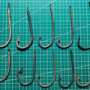 Pode incluir: Uma coleção de dez anzóis de metal individuais dispostos em duas fileiras. Cada anzol apresenta uma haste curva, uma ponta afiada e uma farpa. Os anzóis exibem vários tons de cinza escuro, preto e bronze metálico com sinais de desgaste.