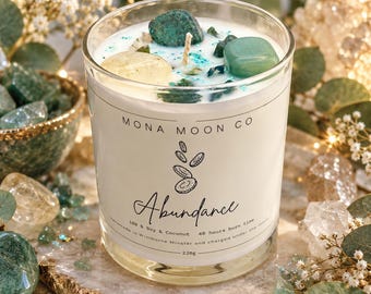 May include: A clear glass candle with white wax topped with green and yellow stones. The label reads MONA MOON CO, Abundance, 100% Soy & Coconut, 40 hours burn time, handmade in Wimborne Minster and charged under the moon, 220g.