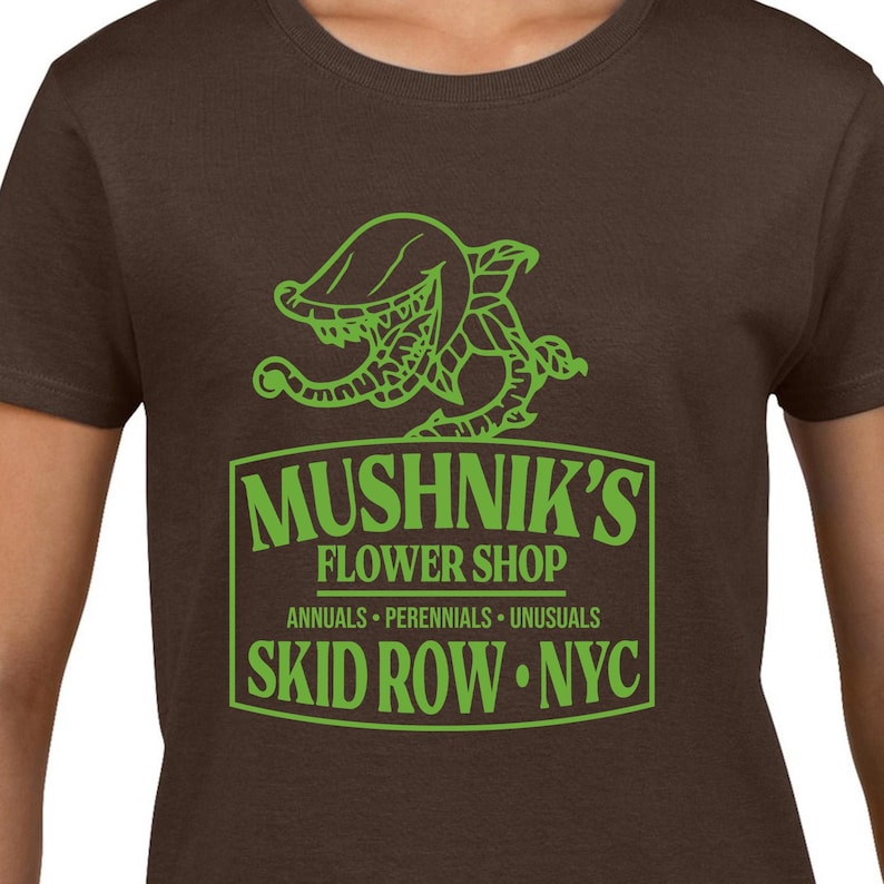 Puede incluir: Una camiseta marr&oacute;n con un gr&aacute;fico verde de una planta carn&iacute;vora y el texto "Mushnik's Flower Shop, Annuals - Perennials - Unusals, Skid Row - NYC."