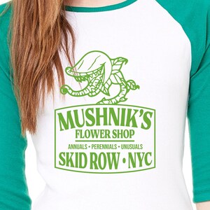 Puede incluir: Una camiseta de b&eacute;isbol blanca y verde con un gr&aacute;fico de una planta carn&iacute;vora y el texto "Mushnik's Flower Shop, Annuals - Perennials - Unusuasls, Skid Row - NYC."