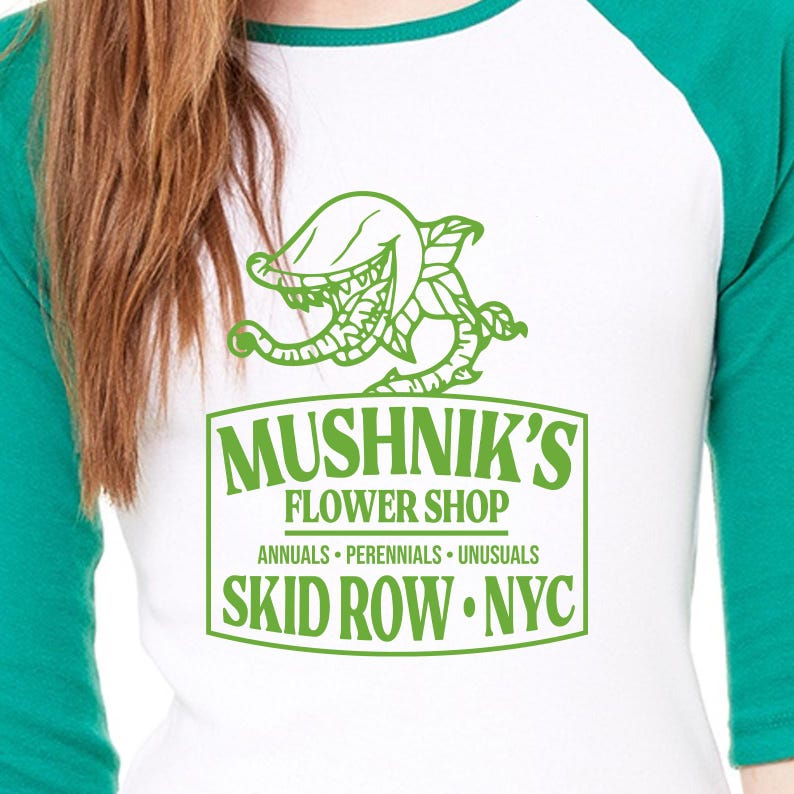 Puede incluir: Una camiseta de b&eacute;isbol blanca y verde con un gr&aacute;fico de una planta carn&iacute;vora y el texto "Mushnik's Flower Shop, Annuals - Perennials - Unusuasls, Skid Row - NYC."