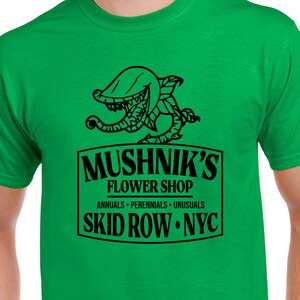 Puede incluir: Una camiseta verde con un gr&aacute;fico negro de una planta carn&iacute;vora y el texto "Mushnik's Flower Shop. Annuals - Perennials - Unusuasl. Skid Row - NYC."