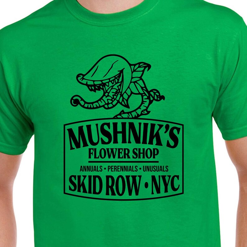 Puede incluir: Una camiseta verde con un gr&aacute;fico negro de una planta carn&iacute;vora y el texto "Mushnik's Flower Shop. Annuals - Perennials - Unusuasl. Skid Row - NYC."