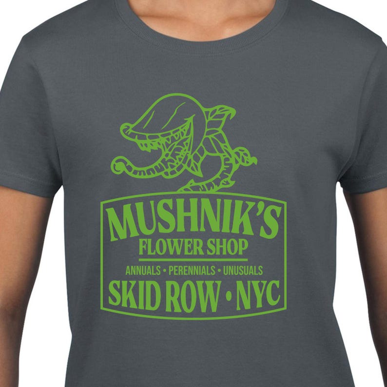 Puede incluir: Una camiseta gris oscuro con un gr&aacute;fico verde de una planta carn&iacute;vora y el texto "Mushnik's Flower Shop, Annuals - Perennials - Unusuasls, Skid Row - NYC."