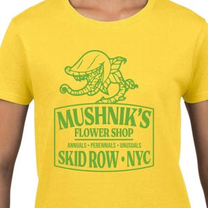 Puede incluir: Camiseta amarilla con un gr&aacute;fico verde de una planta carn&iacute;vora y el texto "Mushnik's Flower Shop. Annuals - Perennials - Unusuasls. Skid Row - NYC."